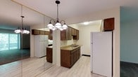 This single-floor, two-bedroom, one-bathroom residence offers a practical and well-organized layout designed for comfortable everyday living. The kitchen serves as a central feature of the space, showcasing rich dark wood cabinetry that provides ample storage across both upper and lower cabinets. Light-toned countertops offer a clean contrast to the darker cabinetry, and a stainless undermount sink is conveniently positioned along the counter run. White appliances, including a full-size refrigerator, are integrated into the kitchen design, complementing the neutral color palette. A decorative pendant chandelier with multiple bell-shaped shades hangs above, adding a warm, inviting focal point to the kitchen area. The flooring throughout features light wood-look planks that lend a clean, contemporary feel to the interior. A floor-to-ceiling mirrored wall panel is positioned adjacent to the kitchen, visually expanding the sense of space and reflecting natural and artificial light throughout the room. The light-toned walls and recessed ceiling details contribute to a bright, airy atmosphere. Overall, the space presents a functional and cohesive design well-suited for everyday living.