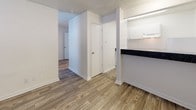 This single-floor unit is designed with a practical, open layout that maximizes the use of available space. Wood-style laminate flooring runs throughout, providing a consistent and clean aesthetic across the living, bedroom, and hallway areas.

The living area is bright and open, featuring a wall-mounted air conditioning unit and a window fitted with horizontal blinds that allows natural light to filter in. Neutral white walls and a textured ceiling contribute to the space's understated, functional character.

A peninsula-style pass-through connects the living area to the kitchen, featuring a dark speckled countertop that contrasts with the white cabinetry above and below. The kitchen is equipped with a dishwasher and ample cabinet storage, offering efficient use of the compact cooking space.

A central hallway links the main living areas to the bedroom and bathroom. The bedroom is a quiet, well-defined room with its own window providing natural light. The full bathroom includes a single-sink vanity with a dark countertop matching the kitchen, white cabinetry, and a tub and shower combination with white tile surround. Wood-style flooring extends into the bathroom, maintaining visual continuity throughout the unit.