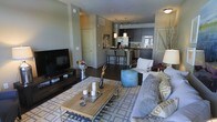This single-floor residence offers a thoughtfully designed open-concept layout that creates a seamless flow between the main living areas. The living room serves as a welcoming focal point, featuring rich hardwood flooring, neutral gray walls, and ample space for comfortable furnishings. A patterned area rug anchors the seating arrangement, which centers around a sleek entertainment console and flat-screen television. Decorative accents, including accent pillows and a teal ottoman, add personality and warmth to the space.

The kitchen is a standout feature, showcasing dark cabinetry that contrasts beautifully with stainless steel appliances and what appears to be dark stone countertops. Pendant lighting above the kitchen bar illuminates the workspace while adding a modern, polished aesthetic. Bar-height seating at the counter peninsula makes the kitchen equally suited for casual dining and entertaining.

Throughout the space, the design maintains a cohesive contemporary palette of grays, neutrals, and warm wood tones. The open floor plan maximizes the sense of space and encourages natural interaction between the kitchen and living areas. Recessed and track lighting supplement the ambiance, ensuring the interiors feel bright and well-appointed at any hour.