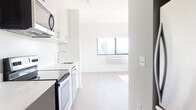 This contemporary single-floor unit showcases a clean, minimalist design throughout, with crisp white walls and wide-plank hardwood flooring that run seamlessly across the living areas and bedrooms. The open-plan kitchen flows directly into the main living space, keeping the layout efficient and connected. The kitchen is outfitted with stainless steel appliances — including a range, built-in microwave, and dishwasher — complemented by white cabinetry with modern hardware and a sleek countertop with an undermount sink. The living area is bathed in natural light from large windows framing impressive city skyline views, reinforcing the elevated position of the unit within the building. Two well-proportioned bedrooms each feature the same hardwood flooring and large windows that invite additional natural light and urban vistas, with wall-unit climate control in each room. The bathroom is finished with dark-tiled flooring, a white vanity with an integrated sink, an illuminated medicine cabinet mirror, and a full soaking tub with an overhead shower. The overall aesthetic is modern and understated, offering a bright, airy atmosphere with a cohesive neutral palette throughout.
