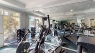 This fitness center is thoughtfully designed to provide a comprehensive workout environment within a single, open floor plan. The space is characterized by elegant tray ceilings with crown molding and recessed lighting, lending a polished, upscale feel that sets it apart from a typical gym setting. Floor-to-ceiling mirrors line the walls, visually expanding the space and allowing users to monitor their form throughout their workout.

The cardio section is well-stocked with multiple treadmills and elliptical machines, arranged to maximize use of the floor space. A dedicated strength training area features a dumbbell rack with a full range of weights alongside a flat workout bench, catering to both beginner and advanced fitness routines. A stationary bike rounds out the equipment selection, offering low-impact cardiovascular options.

Natural light pours in through large, multi-pane windows and glass-paneled doors along one wall, offering pleasant views of the surrounding outdoor greenery. A wall-mounted flat-screen television provides entertainment during workouts. The flooring throughout is durable, cushioned exercise matting in a neutral tone, suited for high-traffic use. Elegant French doors connect the fitness center to an adjacent area, adding an architectural detail that enhances the overall refined aesthetic of the space.