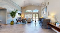 Located at 8001 E 11th Ave in Denver, CO, this single-floor clubhouse offers a thoughtfully designed communal living space that blends modern aesthetics with inviting warmth. The entrance foyer sets an impressive tone, featuring vaulted ceilings, a striking pendant light fixture, and glass-paneled doors that flood the space with natural light. Light-toned wood-style flooring flows seamlessly throughout, enhancing the open and airy atmosphere.

The main living area is generously proportioned, anchored by a sleek gas fireplace framed by a dramatic floor-to-ceiling stone accent wall and a wall-mounted television above. Tall arched windows and expansive glass doors provide sweeping views of the surrounding outdoor area, believed to include a pool, while ceiling fans contribute to year-round comfort. The space is furnished with a mix of sectional sofas, accent chairs, and round side tables, creating multiple comfortable seating zones.

A curated gallery wall displays an eclectic mix of framed artwork, adding a vibrant, creative energy to the room. A marble-topped communal table with pendant lighting defines a dedicated gathering or work area. Lush indoor tropical plants provide natural accents throughout. The clubhouse's cohesive, contemporary design makes it an ideal space for socializing and community gatherings.