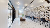 This contemporary office space is thoughtfully designed to support both collaborative and focused work environments. The main office area is anchored by a long communal work surface running along one wall, lined with ergonomic rolling chairs and backed by a striking, large-scale geometric patterned wallcovering in black, white, and gold tones — serving as the space's defining design statement. Gleaming white marble-finish porcelain tile floors run throughout, lending an upscale, professional aesthetic. Track lighting mounted on exposed black rails illuminates the space evenly across the bright white ceiling. A lounge seating arrangement with a modular bench and round side tables offers a more relaxed collaboration zone, while floor-to-ceiling windows along one side flood the interior with natural light and provide views of the outdoors. A glass-partitioned private meeting room, accessible via a sleek black-framed glass wall, accommodates small group discussions with a round table, rolling chairs, a wall-mounted display screen, and a dedicated mini-split HVAC unit. The hallway connects the main office to the entry, finished in the same marble tile and clean white walls, maintaining a cohesive and polished look throughout the entire space.