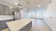 This contemporary single-floor residence showcases a thoughtfully designed open-concept layout that seamlessly connects the kitchen, dining, and living areas. The kitchen is a standout feature, outfitted with sleek gray shaker-style cabinetry extending to the upper walls, crisp white quartz countertops, and a full suite of stainless steel appliances including a refrigerator, range, microwave, and dishwasher. A large center island with a shiplap-style base provides generous prep space and serves as a natural gathering point, accentuated by striking geometric cage pendant lights that hang above. Light wood-look flooring flows throughout the main living areas, lending warmth and continuity to the space. The living area is bright and airy, benefiting from natural light streaming through large windows fitted with horizontal blinds, and is equipped with a ceiling fan for added comfort. Bedrooms feature plush carpeting, ceiling fans, and windows that invite natural light. The bathroom is finished with coordinating gray cabinetry, a white quartz vanity top, a framed mirror, and wood-look tile flooring, maintaining the cohesive modern aesthetic found throughout the residence. Recessed lighting and clean white walls complete the polished, contemporary look of this well-appointed space.