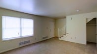 This two-story residential space offers a straightforward and functional layout designed for comfortable everyday living. The main floor features a generously sized living area finished with wall-to-wall carpeting that adds warmth underfoot. A large window along one wall allows natural light to fill the room, creating a bright and open atmosphere. The front entry door is centrally positioned and provides a welcoming point of arrival. Baseboard vents along the lower walls indicate a built-in heating and ventilation system throughout. A classic white-railed staircase anchors one side of the main floor, providing access to the second level. The walls are finished in a neutral, light tone that complements the clean, simple aesthetic of the space. The textured ceiling adds a subtle architectural detail consistent throughout the main level. Overall, the space presents a clean, move-in-ready environment with a neutral palette that offers flexibility for personalization. The two-floor layout provides separation between living and private areas, making the home well-suited for a variety of living arrangements.