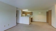 This single-floor unit features an open-concept layout that connects the kitchen and living area into one cohesive space. The kitchen is defined by a white peninsula counter that separates it from the living area while maintaining an open feel. Medium-toned wood cabinetry lines the kitchen walls, complemented by a white refrigerator and stovetop range beneath a built-in ventilation hood. Fluorescent overhead lighting illuminates the kitchen workspace. The living area extends generously from the kitchen, finished with neutral wall-to-wall carpeting that adds warmth underfoot. A pendant light fixture hangs at the far end of the living space, suggesting a designated dining area nearby. The walls throughout are painted in a soft, neutral tone, contributing to a clean and understated ambiance. A textured ceiling is consistent throughout the unit. A hallway visible to the left leads to additional areas of the unit. HVAC venting is integrated into the ceiling, and the overall layout is practical and efficiently organized, making good use of the available space.
