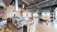 This generously sized single-floor amenity space blends industrial-chic aesthetics with upscale design details to create a dynamic, multi-functional environment. The exposed ceiling reveals structural concrete, painted ductwork, and track lighting, lending an urban loft character throughout. Polished concrete floors run seamlessly across the open layout, tying together each distinct zone.

At the heart of the space is a full demonstration kitchen anchored by a dramatic waterfall-edge marble island with bold geometric tile cladding. Herringbone wood-paneled walls frame the kitchen area, complemented by stainless steel appliances and cylindrical pendant lighting. Adjacent to the kitchen, a dining area features a mix of round and rectangular tables with contemporary seating.

The lounge area is defined by a curved amber leather sectional sofa, sculptural starburst pendant fixtures, and a large-screen television mounted against a gabion stone accent wall. A ping-pong table adds a recreational focal point to the open floor.

Floor-to-ceiling windows wrap the perimeter, flooding the space with natural light and offering sweeping mountain and cityscape views. A set of full-height glass doors opens directly to an outdoor terrace with a pergola-covered patio, extending the living area into the open air. The cohesive design balances comfort, entertainment, and sophistication throughout.