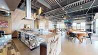 This generously sized single-floor amenity space blends industrial-chic aesthetics with upscale design details to create a dynamic, multi-functional environment. The exposed ceiling reveals structural concrete, painted ductwork, and track lighting, lending an urban loft character throughout. Polished concrete floors run seamlessly across the open layout, tying together each distinct zone.

At the heart of the space is a full demonstration kitchen anchored by a dramatic waterfall-edge marble island with bold geometric tile cladding. Herringbone wood-paneled walls frame the kitchen area, complemented by stainless steel appliances and cylindrical pendant lighting. Adjacent to the kitchen, a dining area features a mix of round and rectangular tables with contemporary seating.

The lounge area is defined by a curved amber leather sectional sofa, sculptural starburst pendant fixtures, and a large-screen television mounted against a gabion stone accent wall. A ping-pong table adds a recreational focal point to the open floor.

Floor-to-ceiling windows wrap the perimeter, flooding the space with natural light and offering sweeping mountain and cityscape views. A set of full-height glass doors opens directly to an outdoor terrace with a pergola-covered patio, extending the living area into the open air. The cohesive design balances comfort, entertainment, and sophistication throughout.
