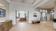 Located at 1 Gradient Court in Weymouth, MA, this single-floor amenity center offers a thoughtfully designed and generously proportioned space for residents to gather, relax, and stay active. The welcoming entry lobby features large-format tile flooring, glass-paneled doors that flood the space with natural light, and a dedicated package locker station for added convenience. A glazed office or leasing area is accessible directly from the lobby.

The centerpiece of the amenity center is a spacious lounge with soaring vaulted ceilings accented by exposed wood beams and shiplap detailing. Warm hardwood-style flooring, a curved sectional sofa, and a vibrant ocean-inspired area rug create an inviting atmosphere. A sleek linear fireplace is flanked by built-in shelving and topped by a mounted flat-screen TV, while a dedicated workspace desk and a marble-topped dining table with floor-to-ceiling drapery complete the social area. Sliding glass doors open to an outdoor patio.

A coastal-themed kitchenette features scalloped ceramic tile backsplash, stainless steel appliances, open display shelving, and a wood-topped bar counter. The expansive fitness center is outfitted with cardio machines, cycling bikes, free weights, resistance equipment, floor-to-ceiling mirrors, and bold circular pendant lighting throughout.