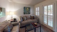 This single-story residence is thoughtfully laid out to maximize functionality across its living spaces. The living room features carpet flooring, a textured ceiling, and a glass-top coffee table, with a door providing access to the exterior and natural light through slatted blinds. It flows directly into the dining area, which is finished with hardwood-style flooring and illuminated by a pendant light overhead. A pass-through window connects the dining space to the kitchen, offering visual openness between the two areas.

The galley-style kitchen is lined on both sides with wood-toned cabinetry and neutral countertops, and is equipped with black appliances including a gas range and refrigerator, as well as a double sink with built-in dishwasher. Hardwood-style flooring continues underfoot, adding warmth to the compact, efficient layout.

The bedroom is carpeted and comfortably sized, featuring a ceiling fan with light fixture, a window with blinds, and ample space for a full furniture arrangement including a dresser and nightstands.

The bathroom includes a vanity with an under-mount sink, wood-style flooring, and a wall-mounted mirror with overhead lighting, offering a clean and practical finish throughout.