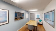 Located at 600 Martin Luther King Jr Blvd in Chapel Hill, NC, this single-room meeting space offers a focused and functional environment well-suited for small group collaboration or professional gatherings. The room features warm hardwood flooring that contrasts pleasantly with the cool blue-gray accent wall, creating an inviting yet professional ambiance. A natural wood conference table takes center stage, surrounded by upholstered gray chairs that comfortably seat a small team. Overhead, a linear fluorescent light fixture provides consistent, even illumination throughout the space. A wall-mounted flat-screen display and a glass-panel whiteboard are positioned on the primary accent wall, equipping the room for presentations and collaborative work sessions. Framed artwork adds a touch of character to the walls. A pair of interior-facing windows with frosted or translucent glass allows borrowed light while maintaining privacy, and a gray paneled door with a glazed insert provides access to the adjoining area. A dark metal bookshelf offers additional storage. The overall design is clean, modern, and practical, making it an efficient venue for meetings and focused work.
