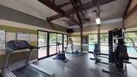Located at 2436 S Holden Rd in Greensboro, North Carolina, this well-equipped fitness center offers a comprehensive workout environment within a single-floor layout. The space is defined by its striking vaulted ceiling with exposed dark wood beams and visible ductwork, lending an open, industrial-rustic character to the room. Horizontal striped walls in shades of olive green, tan, and cream add a bold, energetic aesthetic throughout.

The facility is outfitted with a full range of cardio equipment, including multiple treadmills, an elliptical trainer, and a stationary bike, all positioned on durable rubber flooring for safety and comfort. A multi-station cable and weight machine anchors the strength training area, complemented by a full rack of dumbbells neatly displayed along a mirrored wall. The mirrors effectively expand the sense of space and allow users to monitor their form during exercise.

Natural light fills the room through large windows and glass doors that look out directly onto an outdoor pool and landscaped surroundings, creating an inviting and motivating atmosphere. Wall-mounted televisions and posted workout guides provide added convenience. Ceiling fans help maintain airflow throughout the space. The fitness center is well-maintained and thoughtfully arranged to support a complete workout routine.