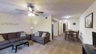 This single-floor residential space offers a welcoming and functional layout designed for comfortable everyday living. The open-concept main area seamlessly connects the living room and dining space, creating a cohesive and practical flow throughout.

The living room is anchored by a plush, dark-toned sofa and loveseat set, accented with patterned throw pillows that add a touch of personality to the neutral color palette. A ceiling fan with an integrated light fixture provides both overhead illumination and air circulation. Soft carpeting underfoot enhances the warmth of the room, while a modest coffee table rounds out the seating area.

Adjacent to the living room, the dining area features a classic wooden table and chair set, ideal for everyday meals. The space transitions smoothly into a hallway leading to additional rooms, with wood-trimmed doorways adding a subtle traditional accent throughout.

The textured ceiling and neutral beige walls contribute to a calm, understated ambiance. Recessed lighting in the hallway ensures the space remains well-lit beyond the main living area. Overall, this unit offers a clean, move-in-ready environment with a straightforward, functional design suited for comfortable residential living.