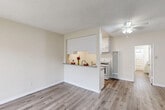 This single-floor unit offers a clean, functional layout unified by wood-look flooring and a neutral warm-beige color palette throughout. The living area is open and well-proportioned, anchored by a ceiling fan with integrated lighting and a textured ceiling. A window near the entry door allows natural light to filter in, with views of surrounding greenery visible through barred windows that add a layer of security.

The kitchen is defined by a half-wall peninsula featuring a granite-style countertop that opens to the living space, creating an efficient pass-through design. Inside, the kitchen is equipped with white cabinetry, a gas range, an over-the-range microwave, a single-basin sink, and a tall floor-to-ceiling ventilation unit. The layout is compact yet practical.

The bedrooms are quiet and simply designed, each finished with the same wood-look flooring. One bedroom features a sliding mirrored closet and ceiling fan, while another includes a window with an outdoor view. A wall-mounted air conditioning unit serves one of the rooms. The bathroom includes a tub-shower combination enclosed by sliding frosted glass doors, white tile surround, and a window above for ventilation.