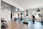 Located at 300 Cityline Ave NE in Atlanta, GA, this well-appointed fitness center offers a comprehensive workout environment within a compact, single-level layout. The space is designed with both function and aesthetics in mind, featuring rich wood-look flooring that runs throughout and crisp white walls that keep the interior feeling bright and open.

A standout architectural detail is the decorative patterned tin ceiling, which covers a portion of the space and adds a distinctive, stylish contrast to the otherwise clean, modern interior. Recessed lighting is embedded throughout, ensuring consistent illumination across the entire floor.

The cardio zone is well-equipped with treadmills, elliptical trainers, a stationary bike, and a stair climber, all positioned along a wall lined with multiple windows that invite natural light and provide views of the surrounding greenery. Ceiling-mounted industrial fans are stationed throughout to maintain comfortable airflow during workouts.

The strength training area includes a multi-station cable machine, a dedicated free weight rack stocked with a full range of dumbbells, and a weight bench. A large wall mirror extends the sense of space and allows for proper form checks during exercise. A wall-mounted air conditioning unit ensures year-round climate control.

Additional amenities include a small shelving unit for personal storage and convenient access to restroom facilities. The space is accessible via a French door entry.