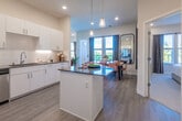 This contemporary single-floor residence at Nova at Queen Valley offers a thoughtfully designed open-concept layout with clean lines and modern finishes throughout. The expansive kitchen serves as the heart of the home, featuring crisp white cabinetry extending to the ceiling, dark quartz countertops, a classic subway tile backsplash, stainless steel appliances including a French door refrigerator and dishwasher, and a central island with pendant lighting overhead. Light wood-look flooring flows seamlessly from the kitchen into the dining area, where large windows and balcony access fill the space with natural light and outdoor views. The living area is bright and airy, with oversized windows framing scenic views of the surrounding landscape. The primary bedroom is a serene retreat featuring plush carpet, a ceiling fan, large windows, and ample natural light. A generously sized walk-in closet with wire shelving provides abundant storage. The bathroom is clean and functional, finished with light flooring, a walk-in shower, and a vanity with a dark countertop. An additional bedroom offers comfortable accommodations with recessed lighting and window views. Throughout the residence, neutral tones, recessed lighting, and high ceilings create a bright, welcoming ambiance.