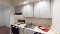 This single-floor residence offers a practical and comfortable layout designed for everyday living. The kitchen is efficiently arranged with ample upper cabinetry featuring classic raised-panel detailing, light-toned countertops, a stainless steel sink, an electric coil range, and a black refrigerator — providing all the essential cooking and storage needs in a compact, well-organized space.

The living area is generously proportioned, featuring rich dark hardwood-style flooring that adds warmth and character throughout. A natural wood entry door anchors the main entrance, and the open flow connects seamlessly to adjacent rooms, including a hallway with a double-door closet offering convenient storage.

The bedrooms are carpeted in neutral tones, creating a soft and quiet atmosphere. Each room provides ample wall space and multiple electrical outlets, offering flexibility for furniture arrangement. At least one bedroom benefits from a window that brings in natural light.

The bathroom is bright and clean, finished with a classic mosaic tile floor in a small-scale grid pattern. It includes a white vanity with under-cabinet storage, a mirrored medicine cabinet with vanity lighting above, and a full toilet — all presented in a crisp, white palette that enhances the sense of cleanliness and space. Overall, the residence combines functional design with comfortable, neutral finishes throughout.