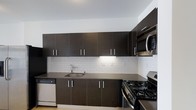 This single-floor space showcases a thoughtfully designed interior with a distinctly modern aesthetic. The kitchen is a standout feature, combining functionality with contemporary style. Dark espresso-toned cabinetry lines both the upper and lower walls, offering generous storage with clean-lined hardware that reinforces the minimalist design language. The countertops are finished in a smooth, dark surface that complements the cabinetry while providing ample workspace. A classic white subway tile backsplash stretches across the full length of the kitchen, adding a crisp contrast to the darker tones and lending a timeless yet modern touch to the space. Stainless steel appliances, including a gas range, over-the-range microwave, and dishwasher, are seamlessly integrated into the layout, completing the professional-grade kitchen setup. The single-basin stainless steel sink is positioned beneath under-cabinet lighting, enhancing both task visibility and ambiance. The overall design of the space is cohesive and polished, with a neutral color palette that creates a clean, uncluttered atmosphere. The layout is efficient and well-organized, making excellent use of the available space throughout the single floor.