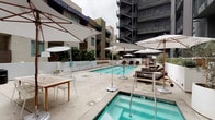 This outdoor amenity space is designed with a contemporary aesthetic, centered around a generously sized lap pool with clear turquoise water and defined lane markings. Adjacent to the pool, a separate spa provides an additional relaxation option, finished in the same clean tile work. The surrounding pool deck is finished in light-toned concrete, offering ample room for lounging and socializing.

Seating throughout the space is thoughtfully arranged, with cushioned chaise lounges positioned along one side of the pool and a casual dining table with chairs on the opposite end. Multiple large market umbrellas are stationed throughout, providing shaded areas across the deck.

The space is enclosed by the facades of the surrounding multi-story buildings, which feature a mix of dark paneling, wood accents, and balconies that frame the courtyard. Landscaping elements include mature olive trees planted in large white cylindrical planters, as well as lush greenery along a low retaining wall, adding a natural softness to the setting.

A standout feature is a large-scale mural painted in bold emerald green and gold tones on a white exterior wall, serving as an artistic focal point. A slatted wood gate with a white metal frame defines the pool entry, contributing to the refined, resort-inspired design language throughout.