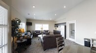 This single-floor space is thoughtfully designed to accommodate both relaxation and functionality in an open, communal setting. The main lounge area is generously proportioned, featuring rich dark-brown leather sofas and armchairs arranged around a round side table, creating an inviting environment for conversation and leisure. Recessed ceiling lighting illuminates the space evenly, complementing the natural light that streams in through multiple large windows along the exterior wall. A wall-mounted flat-screen television adds a modern entertainment element to the lounge.

Adjacent to the seating area, a well-appointed kitchenette offers dark cabinetry, a stainless steel refrigerator, a microwave, and a sink with ample counter space — providing practical amenities for residents or guests. The flooring transitions subtly between patterned carpet in the lounge and smooth hard flooring near the kitchenette, clearly delineating the functional zones within the open layout.

A doorway to the right leads into an additional adjoining room, suggesting further utility or private space. The overall color palette of soft neutral walls, dark furnishings, and warm lighting creates a cohesive, comfortable ambiance that balances both practicality and a welcoming atmosphere.