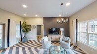 This thoughtfully designed single-floor space makes an immediate impression with its striking chevron-patterned hardwood flooring, which flows seamlessly throughout the open-concept layout and serves as a defining architectural detail. The living area centers around a textured, dark accent wall that anchors a media console and flat-screen television, complemented by a statement chandelier with glass cylinder pendants overhead. A round glass coffee table and paired patterned swivel chairs create an inviting seating arrangement, while large windows allow generous natural light to fill the room.

The kitchen features a substantial island with a quartz countertop and bar stool seating, accented by brass-finish fixtures and pendant lighting. Gray shaker-style cabinetry lines the walls, providing ample storage alongside a clean subway tile backsplash. The adjoining dining nook is positioned beside a large window dressed with bold black-trimmed white curtains, accommodating a round dining table surrounded by upholstered blue chairs. A built-in sideboard with additional cabinetry connects the kitchen and dining areas smoothly. Throughout the space, warm neutral wall tones, recessed lighting, and carefully selected furnishings contribute to a cohesive, contemporary aesthetic.