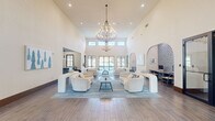 Located at 4600 Portofino Way in West Palm Beach, Florida, this single-floor community clubhouse offers a generously proportioned, thoughtfully designed gathering space with a distinct coastal aesthetic. The centerpiece of the space is a dramatic main living area distinguished by soaring double-height ceilings, clerestory windows that flood the room with natural light, and a striking multi-tiered chandelier that serves as an elegant focal point. The open layout is anchored by plush, curved cream sofas and armchairs arranged around a central coffee table atop a soft blue area rug, all set on wood-look tile flooring that runs throughout. Large glass doors at the rear provide views to the outdoors, enhancing the airy, sun-filled atmosphere. A dedicated business center nook features a built-in desk beneath a dramatic arched alcove adorned with intricate blue-and-white patterned tilework, accommodating multiple workstations. A casual dining area with shiplap-paneled walls offers banquette-style seating with navy velvet benches and warm wood tables, accented by coastal-themed artwork. An additional dining space with grid-pane windows and a modern ceiling fan provides a bright, relaxed setting. The space also includes a kitchen bar area visible from the main lounge, completing this versatile and welcoming clubhouse environment.