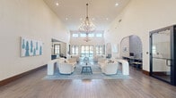 Located at 4600 Portofino Way in West Palm Beach, Florida, this single-floor community clubhouse offers a generously proportioned, thoughtfully designed gathering space with a distinct coastal aesthetic. The centerpiece of the space is a dramatic main living area distinguished by soaring double-height ceilings, clerestory windows that flood the room with natural light, and a striking multi-tiered chandelier that serves as an elegant focal point. The open layout is anchored by plush, curved cream sofas and armchairs arranged around a central coffee table atop a soft blue area rug, all set on wood-look tile flooring that runs throughout. Large glass doors at the rear provide views to the outdoors, enhancing the airy, sun-filled atmosphere. A dedicated business center nook features a built-in desk beneath a dramatic arched alcove adorned with intricate blue-and-white patterned tilework, accommodating multiple workstations. A casual dining area with shiplap-paneled walls offers banquette-style seating with navy velvet benches and warm wood tables, accented by coastal-themed artwork. An additional dining space with grid-pane windows and a modern ceiling fan provides a bright, relaxed setting. The space also includes a kitchen bar area visible from the main lounge, completing this versatile and welcoming clubhouse environment.