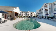 Located at Coventry Commons in Concord, North Carolina, this outdoor amenity space centers around a generously sized, resort-style swimming pool with glistening turquoise water and a gradual entry point. The pool features built-in water fountains that add a dynamic, decorative element to the water surface. Surrounding the pool is a spacious concrete deck furnished with an array of white lounge chairs, outdoor seating groupings with cushioned sofas, and shaded areas beneath blue market umbrellas, providing ample space for relaxation and socializing. A covered pavilion with a ceiling fan and brick detailing anchors one side of the pool area, offering a shaded retreat. A white pergola structure adds further architectural interest and shade on the opposite end of the deck. The pool area is enclosed by a black wrought iron fence with manicured shrubs and seasonal plantings providing additional privacy and greenery. The multi-story residential buildings surrounding the space feature classic architectural details including pitched rooflines, neutral siding, and private balconies. Under clear Carolina skies, the entire outdoor space benefits from abundant natural light, creating a bright, welcoming atmosphere ideal for outdoor recreation and leisure.