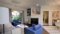 This single-floor residence offers a thoughtfully designed living space that blends comfort with contemporary style. The living room serves as the heart of the home, centered around a classic white-mantled fireplace that adds warmth and elegance to the room. Dark hardwood flooring runs throughout, providing a rich contrast to the light gray walls and white trim. Crown molding accents the ceiling, adding a refined architectural detail to the space.

Natural light pours in through large sliding glass doors and windows dressed with patterned drapes, offering views of lush greenery outside and creating a bright, airy atmosphere. Recessed lighting complements the natural light, ensuring the space is well-illuminated throughout the day and evening.

The living area is generously proportioned, accommodating a full seating arrangement with ease. Bold abstract artwork decorates the walls, lending a modern, curated aesthetic to the room. An open layout connects the living space to adjacent areas of the home, promoting a seamless flow between rooms. The overall ambiance is inviting and well-appointed, balancing clean architectural lines with warm, livable design elements that make this a comfortable and visually appealing space.
