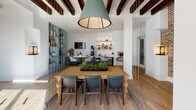 This thoughtfully designed single-floor space blends contemporary style with warm, inviting elements to create a versatile and functional environment. The open layout is anchored by warm wood-tone flooring that flows seamlessly throughout, unifying the distinct functional zones within the space.

A standout lounge area features sage green sofas, pendant lighting with brass accents, and large windows that flood the space with abundant natural light, offering views of the surrounding exterior. Framed botanical artwork adorns the white walls, adding an organic, curated aesthetic.

A central dining or collaborative area sits beneath exposed dark wood ceiling beams and a large teal drum pendant light. A sturdy farmhouse-style wooden table surrounded by upholstered chairs creates an ideal gathering spot, while a floor-to-ceiling teal shelving unit displays decorative accessories nearby.

The kitchenette is well-appointed with sleek gray cabinetry, a white quartz countertop, an undermount sink, a coffee station, microwave, and under-counter refrigerator. A striking herringbone white tile accent wall provides a bold backdrop, complemented by floating wood shelves styled with greenery and pantry essentials.

Overall, the space strikes a balance between modern design and comfortable functionality, making it well-suited for community gathering, collaboration, or shared use.