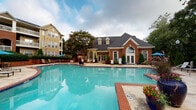 Located at 1003 Glen Forest Dr in Greenville, South Carolina, this well-appointed outdoor amenity area serves as a central gathering space designed for relaxation and recreation. The centerpiece is a generously sized swimming pool with striking blue water, framed by brick edging and a wide surrounding deck that provides ample room for lounging and outdoor seating. Adjacent to the pool, a pergola structure offers shaded relief, sheltering a comfortable outdoor seating arrangement with cushioned wicker furniture — ideal for socializing poolside. A standout feature of the space is the stone-clad built-in grilling station positioned beneath the pergola, making it a functional hub for outdoor cooking and entertaining. Brick knee walls define and border sections of the patio, adding a classic architectural character that complements the traditional brick and siding facade of the clubhouse building visible in the background. Mature trees and well-maintained landscaping, including ornamental shrubs, hanging flower baskets, and potted plants, lend a lush, inviting atmosphere throughout the area. The overall design blends classic Southern architectural elements with practical outdoor amenities, creating a vibrant and community-focused outdoor environment.