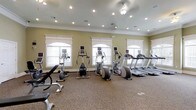 This fitness center offers a bright and welcoming environment for exercise, designed with both functionality and aesthetics in mind. The room features soft sage-green walls accented by crisp white crown molding, giving the space a polished, classic feel. A row of arched windows with decorative trim lines one wall, allowing ample natural light to fill the room and creating an open, airy atmosphere. Recessed lighting and a ceiling fixture provide additional illumination throughout.

The cardio equipment is thoughtfully arranged and includes multiple treadmills, elliptical trainers, and upright exercise bikes, each equipped with individual display screens for tracking workout metrics. The carpeted flooring adds a degree of comfort underfoot, while a weight bench and free weights are also visible, offering options for strength training alongside the cardio machines. The layout provides sufficient space between equipment for ease of movement and safe use. Overall, the fitness center maintains a clean, organized appearance, making it a convenient and comfortable space for residents or guests to pursue their fitness goals.