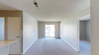 This single-floor space presents a clean, open living area designed with simplicity and functionality in mind. The room is finished with wall-to-wall carpeting in a neutral gray tone, contributing to a calm and understated aesthetic throughout. Smooth, light gray walls are paired with white baseboards, lending the space a clean, cohesive look that serves as a versatile backdrop for a variety of interior styles.

A standout feature of the living area is the sliding glass door positioned along the far wall, which opens to a private balcony and allows natural light to flow into the interior. The balcony railing is visible through the glass, suggesting an outdoor area suitable for relaxation. The natural light entering through this large opening brightens the space considerably, enhancing its open and airy feel.

The ceiling features a textured popcorn finish and includes a centrally placed HVAC vent, indicating a forced-air climate control system. The overall layout is straightforward and open, offering flexibility in furniture arrangement. A secondary door adjacent to the sliding glass door provides additional access or storage options. The space presents a functional, move-in-ready environment with a neutral palette.