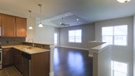 This single-floor residence showcases a thoughtfully designed open-concept layout that seamlessly connects the kitchen and living areas, creating an expansive and inviting atmosphere. The kitchen is equipped with rich dark wood cabinetry, stone countertops, and a functional sink, while stylish pendant lights hang overhead to provide warm, focused illumination. A half-wall partition elegantly defines the transition between the kitchen and the open living space beyond.

The living area is highlighted by a striking vaulted ceiling with a central skylight that draws in abundant natural light, further complemented by a ceiling fan for year-round comfort. Recessed lighting adds a modern finishing touch throughout. Large windows along the far wall flood the interior with daylight and offer views to the exterior, enhancing the airy and open feel of the space.

Underfoot, rich dark hardwood-style flooring runs throughout, adding warmth and visual continuity across the entire floor plan. Neutral gray walls provide a clean, versatile backdrop that complements the home's contemporary aesthetic. The overall design balances functionality and style, making this single-level home a comfortable and visually appealing living environment.