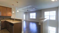 This single-floor residence showcases a thoughtfully designed open-concept layout that seamlessly connects the kitchen and living areas, creating an expansive and inviting atmosphere. The kitchen is equipped with rich dark wood cabinetry, stone countertops, and a functional sink, while stylish pendant lights hang overhead to provide warm, focused illumination. A half-wall partition elegantly defines the transition between the kitchen and the open living space beyond.

The living area is highlighted by a striking vaulted ceiling with a central skylight that draws in abundant natural light, further complemented by a ceiling fan for year-round comfort. Recessed lighting adds a modern finishing touch throughout. Large windows along the far wall flood the interior with daylight and offer views to the exterior, enhancing the airy and open feel of the space.

Underfoot, rich dark hardwood-style flooring runs throughout, adding warmth and visual continuity across the entire floor plan. Neutral gray walls provide a clean, versatile backdrop that complements the home's contemporary aesthetic. The overall design balances functionality and style, making this single-level home a comfortable and visually appealing living environment.