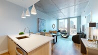 This sophisticated urban residence occupies an elevated floor of a high-rise building, offering breathtaking panoramic city skyline views through dramatic floor-to-ceiling windows. The open-concept living and dining area is anchored by warm hardwood flooring, soft blue accent walls, and a striking exposed concrete ceiling that lends an industrial-chic character to the space. Pendant lighting illuminates the seamless flow between the kitchen and living areas.

The kitchen is thoughtfully designed with crisp white quartz countertops, a subway tile backsplash, two-tone cabinetry in white and natural wood, and a full suite of stainless steel appliances including a gas range, built-in microwave, and French door refrigerator. A spacious island with seating provides an ideal gathering spot.

The living room is bathed in natural light, showcasing a large sectional sofa and glass-top coffee table set against the expansive skyline backdrop. The bedroom continues the refined aesthetic with exposed concrete ceilings, hardwood floors, and a generous window framing city views. The bathroom features large-format tile flooring, a dual-sink vanity with natural wood cabinetry, and a backlit mirror, creating a clean, spa-like atmosphere throughout.