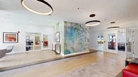 This thoughtfully designed single-floor amenity space offers a generous and versatile layout across multiple interconnected areas. The entry lobby sets a welcoming tone with light wood flooring, a built-in mailroom counter finished in dark gray, and large glass entry doors that invite natural light from the street. Overhead, bold circular pendant fixtures add a contemporary architectural element throughout the common areas.

The central hallway is anchored by a dramatic full-height artistic mural — a vibrant, abstract composition featuring colorful brushstrokes and white branching tree forms — creating a memorable visual focal point. A sleek console table with framed artwork complements this striking installation.

The lounge area features swivel accent chairs, a round coffee table, and floor-to-ceiling windows offering views of the surrounding streetscape. Adjacent to the lounge is a well-appointed kitchen and gathering space with warm wood cabinetry, white quartz countertops, a textured tile backsplash, under-cabinet lighting, and a large island with bar seating beneath matte black pendant lights.

Two dedicated conference rooms accommodate both small and large group meetings, one featuring a bold terracotta accent wall with framed artwork and ample seating around a long communal table. Light wood flooring and clean white walls unify the overall aesthetic throughout.