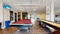 Located at 903-927 10th Ave SW in Calgary, Alberta, this generously sized common room offers a well-appointed, multi-functional amenity space designed for recreation and socializing. The open-concept layout flows across a single floor, unified by warm hardwood-style flooring and a striking coffered ceiling with recessed lighting that adds architectural depth and a sense of elevated refinement throughout.

The recreation zone is a central highlight, featuring a full-size pool table with a bold red felt surface, a shuffleboard table, and a ping pong table — providing a diverse range of gaming options. A dedicated dining and casual seating area offers stylish bentwood chairs in teal paired with round bistro tables, ideal for informal gatherings. Adjacent to this is a comfortable lounge zone furnished with vibrant red armchairs, a striped sofa, and a glass coffee table, bathed in natural light from floor-to-ceiling windows dressed with sheer curtains.

A colorful stained-glass-style panel installation serves as a distinctive artistic focal point, lending character to the space. Framed artwork is displayed throughout, adding a curated gallery feel. Large glazed doors provide direct access to an outdoor terrace, further enhancing the connection to the surrounding cityscape. The space is well-suited for both active recreation and relaxed social engagement.