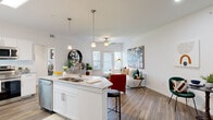 This thoughtfully designed single-floor residence offers a seamless open-concept layout that connects the kitchen and living area into one bright, welcoming space. The kitchen is outfitted with crisp white shaker cabinetry, granite-style countertops, stainless steel appliances including a refrigerator, range, and microwave, and an island with a double-basin sink and pendant lighting overhead. Wood-look flooring runs throughout the entire residence, providing a warm, cohesive aesthetic.

The living area benefits from natural light streaming through multiple windows and a glass-paned door, complemented by a ceiling fan. The open flow between spaces creates a generous sense of room ideal for both relaxing and entertaining.

The bedroom is a comfortable retreat, featuring a ceiling fan, a window with views of greenery, and ample wall space. Clean white walls and wood-look flooring carry through here as well. An additional room visible in the floor plan provides flexible use as a second bedroom or den.

The bathroom is clean and modern, featuring white cabinetry with granite-style countertops, a walk-in glass-enclosed shower with chrome fixtures, and wood-look tile flooring. A dedicated laundry area with washer and dryer connections rounds out the functional amenities of this well-appointed residence.