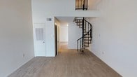 This two-floor space makes a strong first impression with its dramatic double-height ceilings and an eye-catching spiral staircase with sleek black metal railings that serves as a central architectural focal point. The open main living area is generously proportioned, finished with wall-to-wall neutral carpeting that adds warmth and comfort underfoot. Crisp white walls throughout reflect light effectively, contributing to a clean and airy ambiance. The spiral staircase connects to an upper-level loft or mezzanine area, visible from below with matching black metal railings that maintain a cohesive design language across both floors. Adjacent to the main living space, a hallway leads to additional rooms, suggesting a functional and well-organized floor plan. Practical elements such as a utility access panel and HVAC vent are neatly integrated into the walls. The overall design balances open, voluminous living areas with defined private spaces, making it a versatile and visually engaging two-story layout suited to a variety of uses.