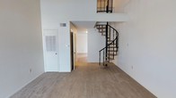 This two-floor space makes a strong first impression with its dramatic double-height ceilings and an eye-catching spiral staircase with sleek black metal railings that serves as a central architectural focal point. The open main living area is generously proportioned, finished with wall-to-wall neutral carpeting that adds warmth and comfort underfoot. Crisp white walls throughout reflect light effectively, contributing to a clean and airy ambiance. The spiral staircase connects to an upper-level loft or mezzanine area, visible from below with matching black metal railings that maintain a cohesive design language across both floors. Adjacent to the main living space, a hallway leads to additional rooms, suggesting a functional and well-organized floor plan. Practical elements such as a utility access panel and HVAC vent are neatly integrated into the walls. The overall design balances open, voluminous living areas with defined private spaces, making it a versatile and visually engaging two-story layout suited to a variety of uses.