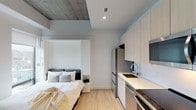 This thoughtfully designed studio unit offers a compact yet highly functional single-floor layout that maximizes every square foot through intelligent use of space. The open-plan living and sleeping area centers around a built-in Murphy bed system, which folds seamlessly into the wall to reveal flexible living space when not in use. A large window flanks the sleeping area, flooding the room with natural light and framing panoramic city views of the surrounding urban neighborhood.

The kitchen is well-appointed with light wood flat-front cabinetry extending to the ceiling for ample storage, stainless steel appliances including a refrigerator, range, dishwasher, and microwave, along with a sleek countertop and undermount sink. Light-toned hardwood flooring runs throughout the main living area, contributing to the unit's bright and airy aesthetic.

A dedicated workspace with a built-in desk and seating provides an area for focused work or study. The unit also includes a stacked in-unit washer and dryer, adding a high level of convenience. The bathroom features a clean, minimalist design with white tile and a glass-enclosed rainfall shower. Exposed concrete ceiling elements in select areas lend an industrial character that contrasts beautifully with the otherwise crisp, modern interior palette.