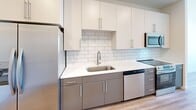 Located at 1803 S 46th St in Rogers, Arkansas, this thoughtfully designed one-bedroom, one-bathroom unit offers a clean, modern aesthetic across its single-floor layout. The galley-style kitchen is a standout feature, equipped with stainless steel appliances including a refrigerator, range, microwave, and dishwasher. White upper cabinetry pairs with gray lower cabinets, complemented by a white subway tile backsplash, white quartz countertops, and warm hardwood-style flooring, creating a contemporary two-tone design. The bedroom is finished with soft carpeting, a ceiling fan, and a window that allows natural light and exterior views to fill the room. The bathroom features hardwood-style flooring, a white subway tile walk-in shower with brushed nickel fixtures, a single-sink vanity with white countertop, and a mirror. A dedicated laundry area houses a stacked front-loading washer and dryer unit, adding everyday convenience. Throughout the space, neutral white walls, recessed lighting, and consistent flooring choices create a cohesive, bright, and functional living environment well suited for comfortable single-occupant living.