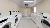 Located at 3817 George Washington Hwy in Portsmouth, Virginia, this single-floor community laundry facility is designed to serve residents with efficiency and convenience. The space is organized with rows of front-loading washing machines and top-loading dryers lining both sides of the room, providing ample capacity for simultaneous use by multiple residents. The interior features light-colored walls that contribute to a clean and open atmosphere, complemented by bright fluorescent overhead lighting that ensures excellent visibility throughout. The flooring consists of durable, easy-to-maintain tile, well-suited for a high-traffic utility environment. A small wooden shelving unit positioned centrally along one wall offers a practical surface for folding or setting down laundry items. Informational and safety notices are posted on the walls, keeping users informed of facility rules and procedures. A bulletin board near the entry door provides a space for community communications. Ventilation is handled through ceiling-mounted HVAC registers, maintaining air circulation throughout the room. A standard six-panel door provides access to and from the facility. The overall layout is straightforward and functional, prioritizing ease of use and efficient flow, making this laundry facility a practical and well-maintained amenity.