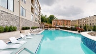 Located at 4021 Ridge Ave in Philadelphia's East Falls neighborhood, this generously sized outdoor amenity space offers a resort-inspired experience within a modern residential community. The centerpiece is a large, shimmering pool with vivid blue tile accents, flanked by an extensive sun deck lined with contemporary lounge chairs and in-pool tanning ledges, perfect for relaxing in the open air.

Surrounding the pool, the space is thoughtfully designed with multiple distinct zones. A covered outdoor kitchen area features stainless steel grills set against a striking natural stone accent wall, sheltered beneath a dramatic curved black steel pergola equipped with ceiling fans — ideal for al fresco dining and entertaining. Freestanding canopy cabanas are positioned poolside, offering shaded retreats with daybed-style furnishings.

Raised planter beds filled with manicured shrubs and seasonal greenery line the perimeter, adding lush, natural texture to the hardscaped surroundings. Black steel fencing defines the boundary of the space while maintaining open views of the adjacent wooded hillside and neighboring brick buildings. The multi-story residential building wraps the courtyard, with balconies overlooking the amenity area. The overall ambiance is polished, functional, and resort-like.