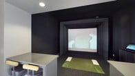 This dedicated golf simulator room is purpose-built for an immersive indoor golf experience. The space is finished with matte black walls and ceiling, creating a dark, focused environment that enhances the visibility and contrast of the large projection screen mounted at the far end of the room. The screen is framed by a substantial black enclosure that doubles as a protective barrier, ensuring a clean and professional setup.

At the center of the room, a synthetic turf hitting mat with designated tee positions provides a realistic feel underfoot, simulating the sensation of playing on an actual course. The dark carpet flooring surrounding the mat further absorbs light and contributes to the room's immersive atmosphere. Recessed ceiling lighting provides adequate illumination without causing glare on the screen.

A counter or cabinet along one side of the room offers a convenient surface for equipment, beverages, or controls. The overall design is streamlined and utilitarian, with every element serving the primary function of delivering a high-quality, distraction-free golf simulation experience suitable for practice or leisure play.