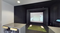 This dedicated golf simulator room is purpose-built for an immersive indoor golf experience. The space is finished with matte black walls and ceiling, creating a dark, focused environment that enhances the visibility and contrast of the large projection screen mounted at the far end of the room. The screen is framed by a substantial black enclosure that doubles as a protective barrier, ensuring a clean and professional setup.

At the center of the room, a synthetic turf hitting mat with designated tee positions provides a realistic feel underfoot, simulating the sensation of playing on an actual course. The dark carpet flooring surrounding the mat further absorbs light and contributes to the room's immersive atmosphere. Recessed ceiling lighting provides adequate illumination without causing glare on the screen.

A counter or cabinet along one side of the room offers a convenient surface for equipment, beverages, or controls. The overall design is streamlined and utilitarian, with every element serving the primary function of delivering a high-quality, distraction-free golf simulation experience suitable for practice or leisure play.