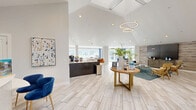 Located at 5000 S Lincoln Trace Ave SE in Smyrna, Georgia, this single-floor community space offers a thoughtfully designed interior that combines modern aesthetics with comfortable, functional living. The space is anchored by a dramatic vaulted ceiling in the main living area, which rises to impressive heights and is adorned with a contemporary circular chandelier, lending a sense of grandeur to the open floor plan. Large-format light-toned tile flooring runs throughout, contributing to a clean, cohesive look that reflects natural light beautifully. Floor-to-ceiling windows and multiple window groupings line the walls, flooding every area with abundant natural light and offering lush outdoor views of surrounding greenery. The living area is generously proportioned, featuring distinct lounge and workstation zones, with a textured wood-paneled accent wall serving as a standout architectural detail. A large flat-screen display and herringbone-front media console anchor one end of the room, while velvet blue seating accents add a pop of color throughout. The kitchen is sleek and modern, outfitted with dark cabinetry, stainless steel appliances, and light countertops, complemented by a windowed wall that keeps the space bright and airy. Overall, the space exudes a polished, welcoming ambiance ideal for both collaboration and relaxation.