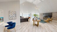 Located at 5000 S Lincoln Trace Ave SE in Smyrna, Georgia, this single-floor community space offers a thoughtfully designed interior that combines modern aesthetics with comfortable, functional living. The space is anchored by a dramatic vaulted ceiling in the main living area, which rises to impressive heights and is adorned with a contemporary circular chandelier, lending a sense of grandeur to the open floor plan. Large-format light-toned tile flooring runs throughout, contributing to a clean, cohesive look that reflects natural light beautifully. Floor-to-ceiling windows and multiple window groupings line the walls, flooding every area with abundant natural light and offering lush outdoor views of surrounding greenery. The living area is generously proportioned, featuring distinct lounge and workstation zones, with a textured wood-paneled accent wall serving as a standout architectural detail. A large flat-screen display and herringbone-front media console anchor one end of the room, while velvet blue seating accents add a pop of color throughout. The kitchen is sleek and modern, outfitted with dark cabinetry, stainless steel appliances, and light countertops, complemented by a windowed wall that keeps the space bright and airy. Overall, the space exudes a polished, welcoming ambiance ideal for both collaboration and relaxation.
