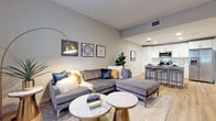 This single-floor residence offers a thoughtfully designed layout that balances functionality with contemporary style. Light hardwood flooring flows throughout, creating a warm and cohesive foundation across each space.

The open-concept kitchen is a standout feature, outfitted with crisp white shaker cabinetry, stainless steel appliances including a range, microwave, and French door refrigerator, and a light quartz-topped island with seating for multiple guests. Recessed lighting above highlights the clean, modern aesthetic.

The living area is bright and welcoming, benefiting from natural light through large windows dressed with floor-length draping. Neutral gray walls provide a versatile backdrop, and the open sightline to the kitchen enhances the sense of space.

The bedroom is generously proportioned, accommodating a full suite of furniture comfortably. A ceiling fan, natural light from windows with shuttered treatments, and a calm, neutral color palette contribute to a restful atmosphere.

The bathroom features a full soaking tub surrounded by white subway tile, offering a clean and timeless finish. Throughout the residence, the design maintains a consistent modern sensibility with neutral tones and quality finishes.